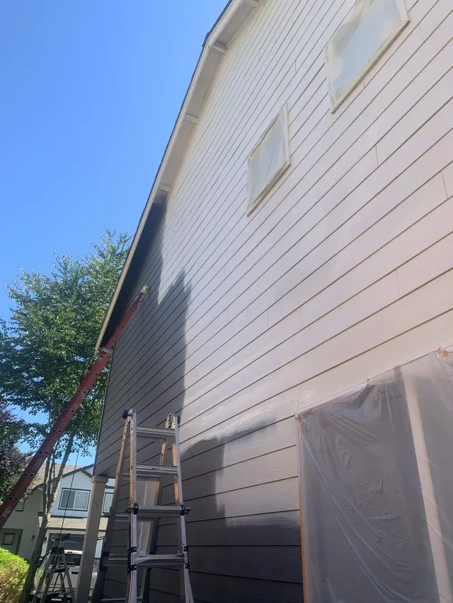 Windows and doors fully masked in plastic while siding is sprayed, with ladders in place